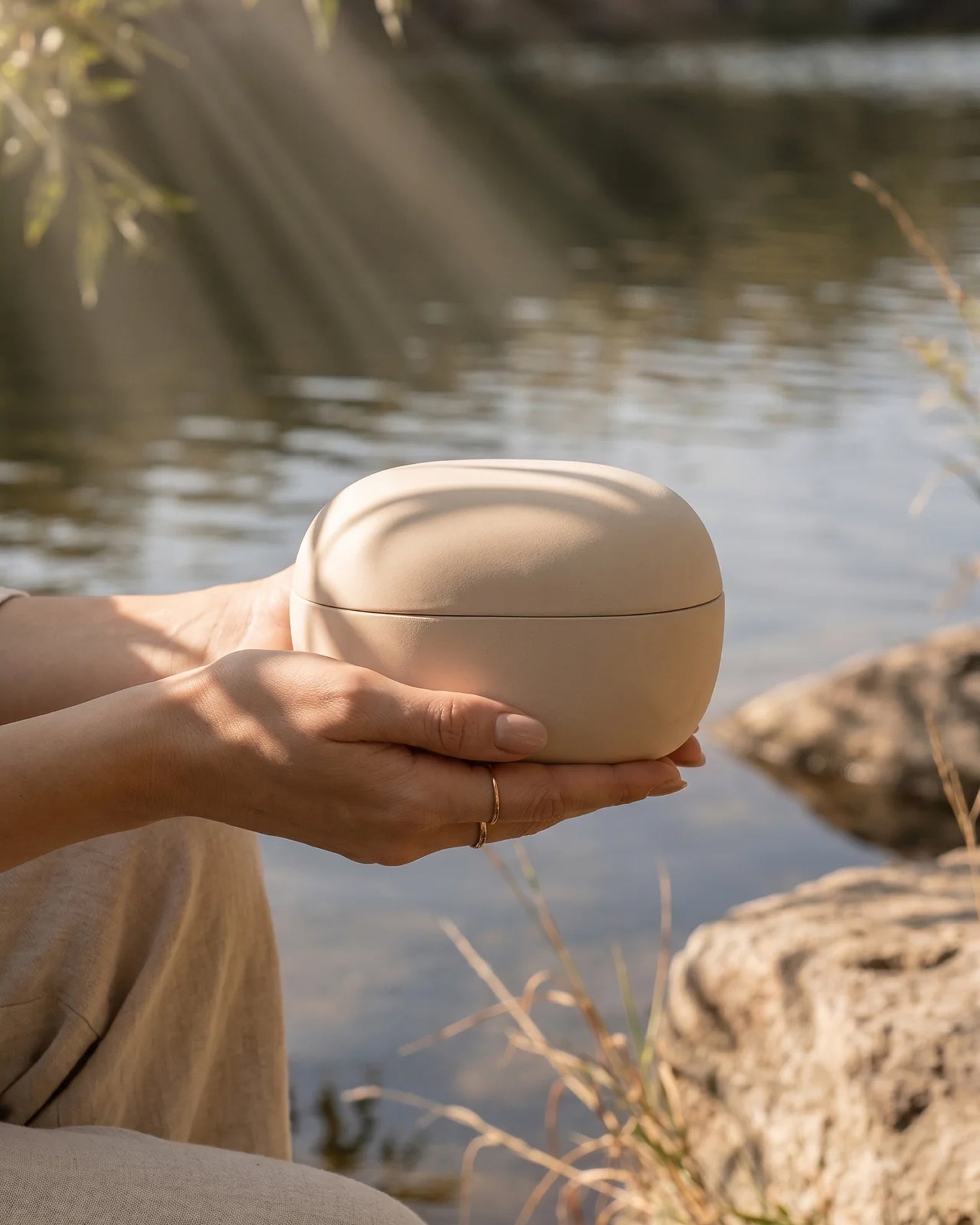 Eco-friendly cremation urn held in hands near water — natural clay biodegradable urn for water scattering