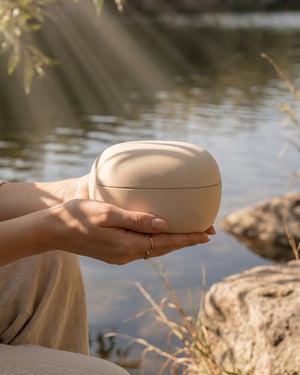 Eco-friendly cremation urn held in hands near water — natural clay biodegradable urn for water scattering